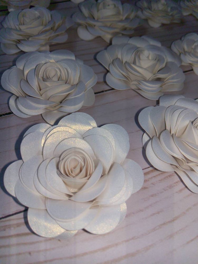 May include: A collection of white paper flowers with a subtle shimmer. The flowers are layered and have a delicate, feminine appearance.