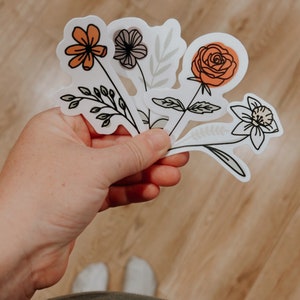 May include: A set of five floral stickers featuring different flower designs in orange, white, and gray. The stickers are on a white background and have a hand-drawn style.