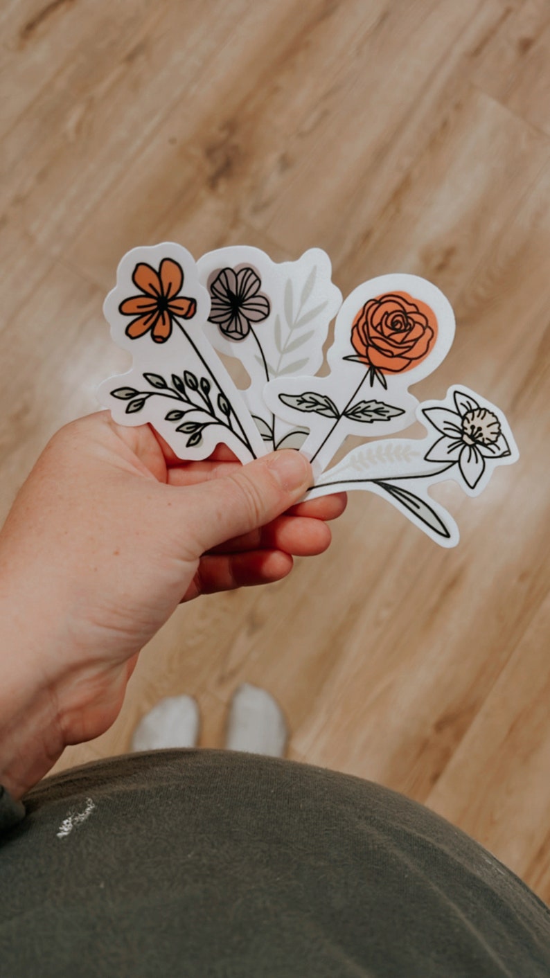 May include: A set of five floral stickers featuring different flower designs in orange, white, and gray. The stickers are on a white background and have a hand-drawn style.