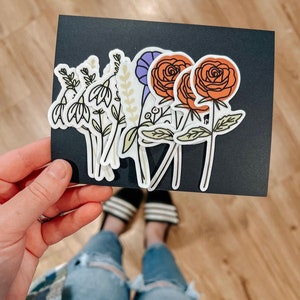 May include: A set of six floral stickers featuring various flowers, including a purple flower, a white flower with green leaves, and two orange roses with green leaves.