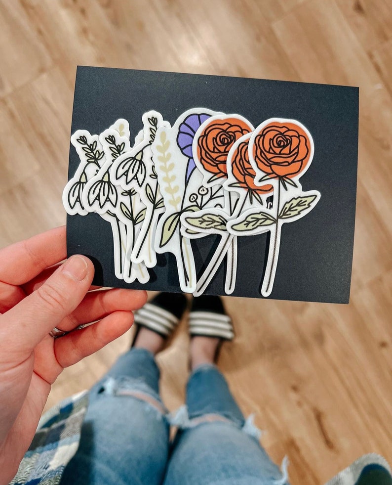 May include: A set of six floral stickers featuring various flowers, including a purple flower, a white flower with green leaves, and two orange roses with green leaves.