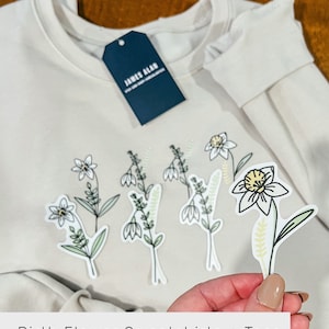May include: A cream-colored sweatshirt with a navy blue tag that says "JAMES ALAN" and several white, green, and yellow flower stickers. The text "Birth Flower Sweatshirts + Tees available in another listing." is at the bottom of the image.