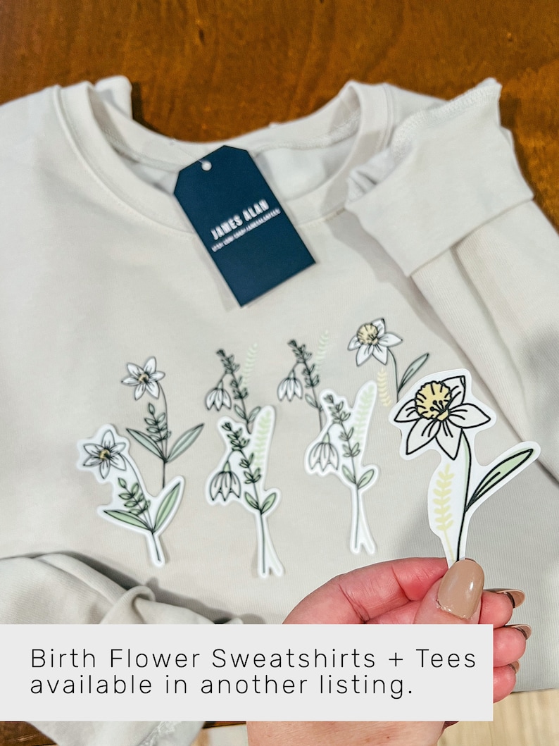 May include: A cream-colored sweatshirt with a navy blue tag that says "JAMES ALAN" and several white, green, and yellow flower stickers. The text "Birth Flower Sweatshirts + Tees available in another listing." is at the bottom of the image.