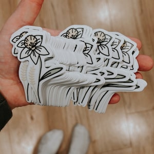 May include: A stack of white stickers with a black outline of a daffodil flower with a brown center. The flower has a stem with two brown leaves and two brown wheat stalks.