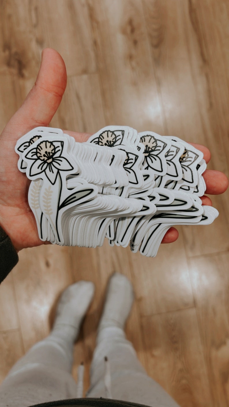 May include: A stack of white stickers with a black outline of a daffodil flower with a brown center. The flower has a stem with two brown leaves and two brown wheat stalks.