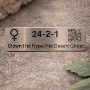 May include: A rectangular, brushed metal tag with black text and a QR code. The tag features a female symbol, the numbers "24-2-1", and the words "Clown Het Hypo Het Desert Ghost". The background is a textured, neutral surface.