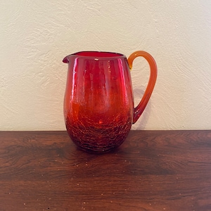 May include: A red glass pitcher with a textured surface and an amber handle. The pitcher is on a wooden surface.