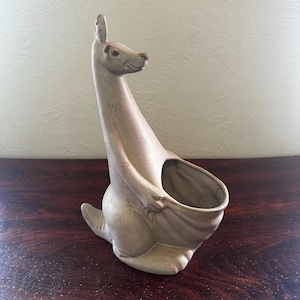May include: A ceramic kangaroo figurine in a speckled beige finish. The kangaroo is depicted in a standing pose with a pouch-like opening in its belly. The figurine has a long neck and a detailed face, and is likely a decorative item.