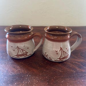 May include: Two ceramic mugs with a brown rim and handle, and a light gray body. Each mug features a brown sailboat design with the word "lake" below it. The mugs are on a wooden surface.