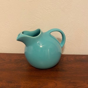 May include: A turquoise ceramic pitcher with a rounded body and a curved spout. The pitcher has a handle on the side.