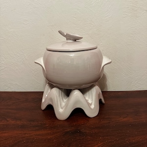 May include: A white ceramic serving dish with a lid and a wavy base. The dish has a delicate, floral design on the lid.