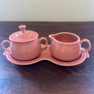 May include: A vintage pink ceramic sugar bowl with a lid and a creamer, both with handles, sit on a matching tray. The set has a classic, retro design and is likely from the mid-20th century. The pieces are a solid, muted pink color.