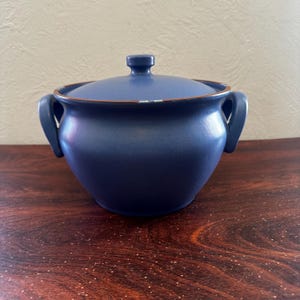 May include: A blue ceramic serving dish with two handles and a lid. The dish has a slightly rounded shape and a brown rim.