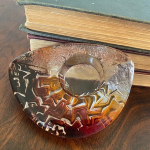 May include: A clear glass candle holder with a textured, abstract design in shades of brown, orange, red, and white. The holder has a round opening in the center for a candle.