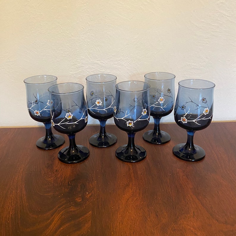 May include: Set of six blue glass wine glasses with white floral designs. The glasses have a clear base and a wide bowl.
