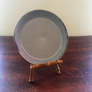 May include: A gray ceramic plate with a dark gray rim, displayed on a small bamboo stand. The plate has a circular design and is set against a dark wood surface. The bamboo stand has a natural, light brown color.
