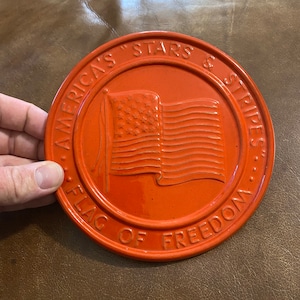 May include: Orange ceramic trivet with a raised image of the American flag and the text "America's "Stars & Stripes" Flag of Freedom".