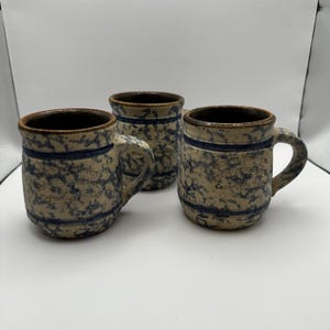 May include: Three antique stoneware mugs with handles. The mugs have a speckled beige and blue design with a dark brown rim and a blue band. The mugs have text on the front. The mugs are likely for drinking beverages.