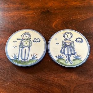 May include: Two ceramic coasters with blue and white designs. One coaster features a cartoon boy holding a rake and the other features a cartoon girl wearing a blue dress.