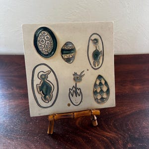 May include: A square ceramic tile with embossed designs of eggs and baby birds in oval frames. The designs are in shades of teal and gray, set against a cream-colored background. The tile is displayed on a small bamboo easel.
