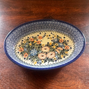 May include: Oval ceramic serving dish with a blue rim and floral design. The rim features a detailed blue pattern. The center of the dish displays a bouquet of flowers in shades of orange, blue, and yellow. The dish is set on a dark wooden surface.