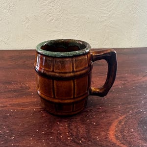 May include: A brown ceramic mug shaped like a barrel with a green glazed rim. The mug has a handle and is sitting on a wooden surface.