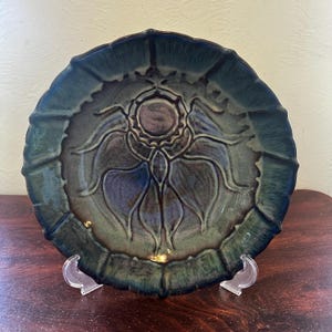 May include: A ceramic plate with a floral design. The plate is a deep teal color with a darker center and a scalloped edge. The floral design is raised and has a textured appearance. The plate is displayed on a clear stand.