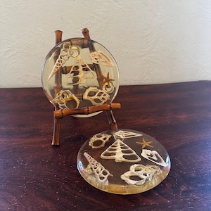 May include: Two round clear resin coasters with embedded seashells and a starfish. The coasters are displayed on a small wooden easel.