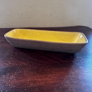 May include: A rectangular ceramic dish with a yellow interior and a textured, brown exterior. The dish has a simple, yet elegant design, perfect for serving food or as a decorative piece. The dish is approximately 12 inches long.