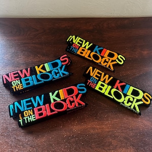 May include: Four embroidered patches with the text "NEW KIDS ON THE BLOCK" in vibrant colors. The patches are black with the text in pink, blue, yellow, orange, and green. The patches are arranged on a dark brown wooden surface.