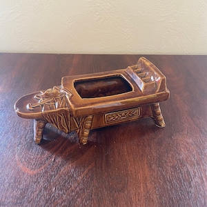 May include: Brown ceramic ashtray in the shape of a wheelbarrow. The ashtray has a rectangular opening for ashes and a detailed design of a wheelbarrow with a decorative pattern on the side. The ashtray is supported by four small legs.