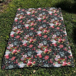 May include: A black floral picnic blanket with a large-scale floral pattern in shades of pink, red, blue, and green. The blanket is laid out on a grassy surface.