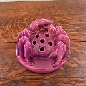 May include: A ceramic flower frog in a deep magenta color, shaped like a crab. The crab has multiple holes for arranging stems and a circular base. The crab's claws and body are detailed, adding to its unique design.
