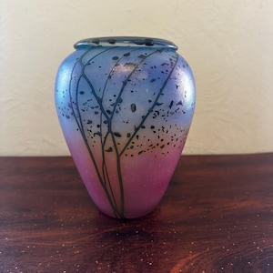 May include: A decorative glass vase with a rounded shape and a wide opening. The vase features a gradient of colors, transitioning from pink at the base to blue at the top. Dark, branch-like patterns are painted on the surface.