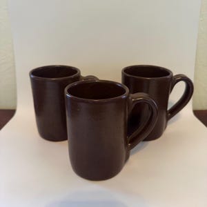 May include: Three brown ceramic mugs with handles. One mug is centered, with two others on either side. The mugs have a simple, cylindrical shape and a smooth, matte finish. The mugs are suitable for coffee, tea, or other beverages.