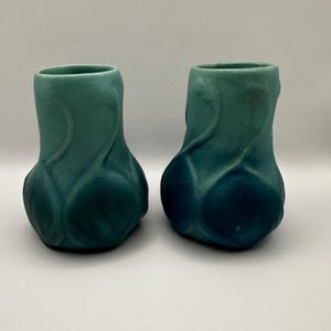 May include: Two teal ceramic vases with a textured, organic design. The vases have a bulbous base that tapers to a narrow opening. The design features swirling, leaf-like patterns and a gradient of teal to deep blue.