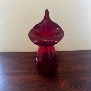 May include: A red glass vase with a wide, flared opening and a narrow neck. The vase has a textured surface and a unique, organic shape.