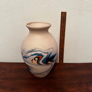 May include: A beige ceramic vase with a wide body and narrow neck. The vase features a swirling pattern of blue, orange, and black colors. A wooden ruler is placed next to the vase. The vase is approximately 12 inches tall.