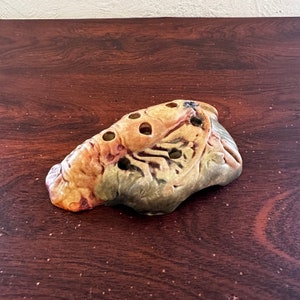 May include: A ceramic flower frog in the shape of a stylized fish with a brown, green, and yellow glaze. The frog has multiple holes for holding flower stems.