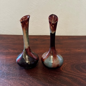 May include: Two small ceramic vases with a brown, blue, and white glaze. The vases have a narrow neck and a rounded base.