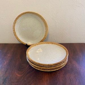 May include: A set of six off-white ceramic plates with speckled surfaces and a brown rim. One plate is propped up, while the others are stacked on a dark wooden surface. The plates have a rustic, handmade appearance.