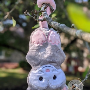 May include: A gray and white plush toy with pink ears and paws hangs upside down from a tree branch. The toy has embroidered eyes and a pink nose.