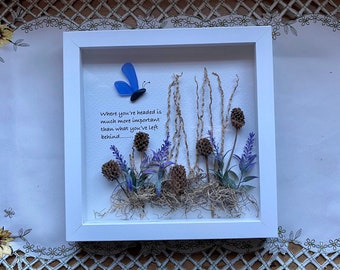May include: A white framed shadow box with a blue butterfly, dried flowers, and the quote "Where you're headed is much more important than what you've left behind..."