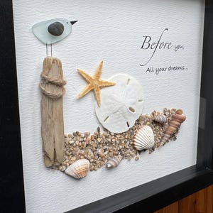 May include: Framed artwork with a seaside theme. A glass bird sits on a wooden post wrapped with rope. A starfish, sand dollar, and seashells are arranged on a bed of pebbles. The text reads "Before you, All your dreams..."