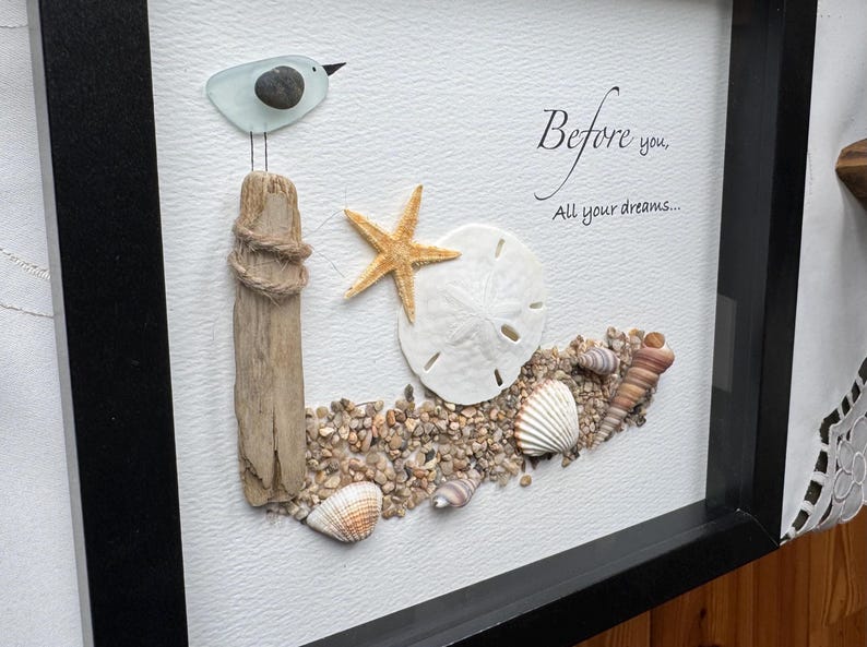 May include: Framed artwork with a seaside theme. A glass bird sits on a wooden post wrapped with rope. A starfish, sand dollar, and seashells are arranged on a bed of pebbles. The text reads "Before you, All your dreams..."