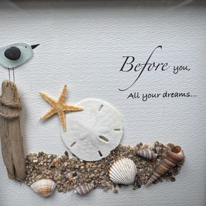 May include: Framed artwork depicting a beach scene with a glass bird on driftwood. A starfish, sand dollar, and seashells are arranged on pebbles. The text "Before you, All your dreams..." is written in elegant script.