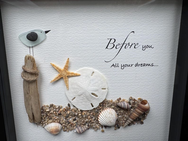 May include: Framed artwork depicting a beach scene with a glass bird on driftwood. A starfish, sand dollar, and seashells are arranged on pebbles. The text "Before you, All your dreams..." is written in elegant script.