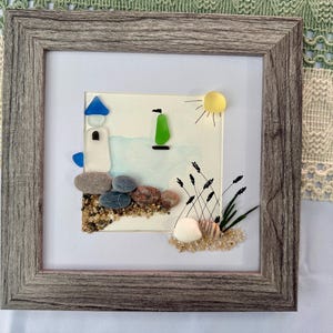 May include: Framed artwork featuring a coastal scene. A lighthouse made of white and blue glass sits near a beach made of sand and pebbles. A green glass sailboat sails on the blue water under a yellow sun. The frame is made of gray wood.