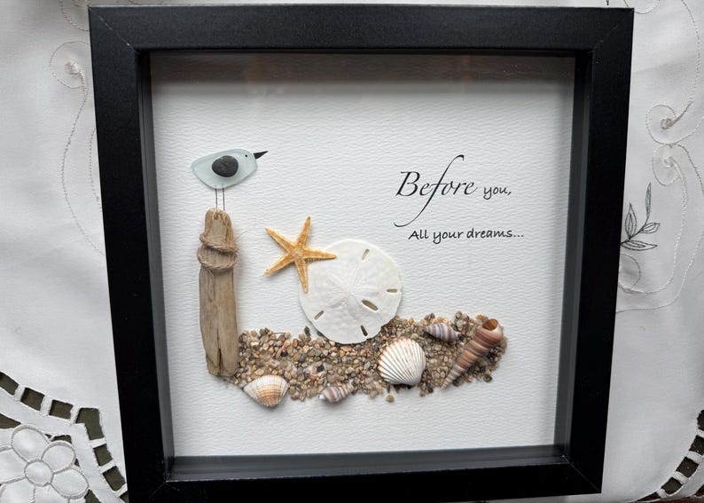 May include: Framed artwork depicting a beach scene with a seagull, driftwood, starfish, sand dollar, and seashells. The text "Before you, All your dreams..." is written in script. The frame is black.