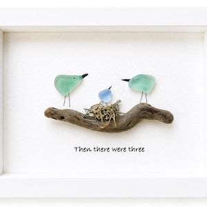 May include: A framed wall art piece featuring three birds crafted from sea glass and wire perched on a piece of driftwood. Two larger light green birds flank a smaller blue bird sitting in a nest made of twigs. The text reads, Then there were three.